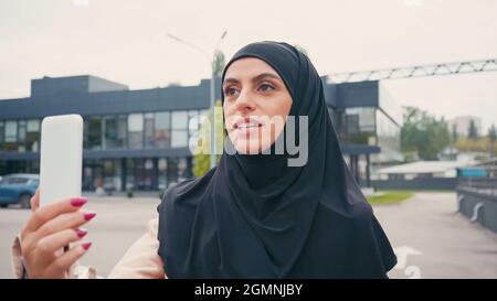 Junge muslimische Frau im Hijab, die während eines Videoanrufs auf der städtischen Straße spricht Stockfoto