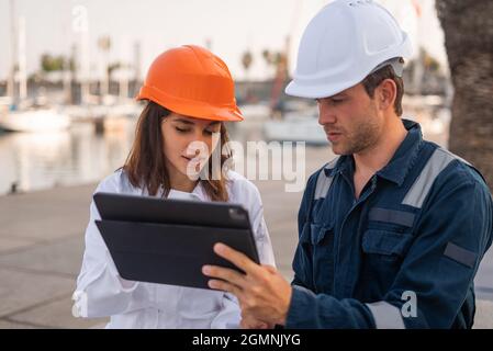 Seriöse männliche Ingenieurin und weibliche Inspektorin in Hardhats diskutieren Informationen auf Tablette während des Treffens im Seehafen Stockfoto