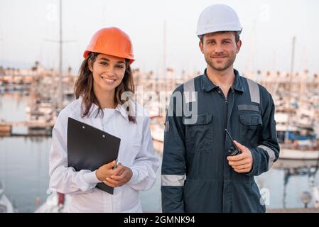 Lächelnde, junge Hafenmitarbeiter in Arbeitskleidung und Helmen, die die Kamera betrachten, während sie mit Clipboard und Walkie Talkie am Pier in der Nähe von festgetäuten Schiffen arbeiten Stockfoto