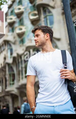Niedriger Winkel des selbstbewussten jungen bärtigen männlichen Touristen in weißem T-Shirt mit Rucksack, der mit der Hand in der Tasche steht und am Sommertag gegen das alte Stadtgebäude blickt Stockfoto