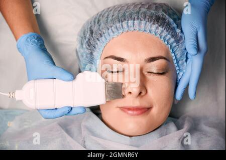 Nahaufnahme der Hände der Kosmetikerin mit Ultraschallschrubber beim Reinigen des weiblichen Gesichts der Kundin. Frau in Einweg-Duschhut mit Tiefenreinigung Hautpflege Verfahren. Konzept der Schönheit, Hautpflege Behandlung. Stockfoto