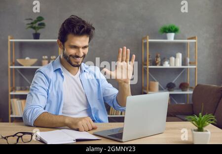 Glücklicher Mann, der mit einem Laptop am Schreibtisch saß, lächelte und mit der Hand am Bildschirm winkte Stockfoto