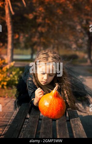 Halloween. Ein süßes Mädchen mit langen Haaren liegt auf der Bank im Park und zieht ein lustiges Gesicht zu Kürbis. Stockfoto