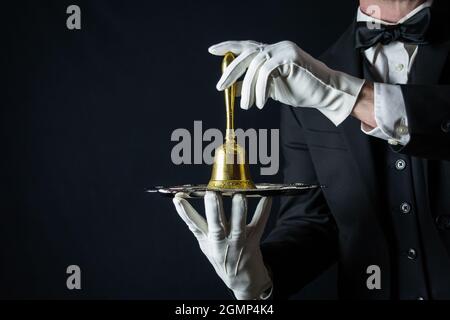 Butler oder Kellner in Dunkelanzug und weißen Handschuhen mit Goldglocke auf Silbertablett. Konzept der Dienstleistungsbranche und der professionellen Gastfreundschaft. Stockfoto