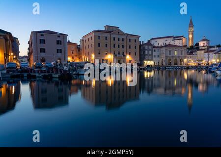 Piran Hafen am Abend bei Blue hur mit Fischerbooten und beleuchteten Gebäuden st. george Kirchturm. Stockfoto