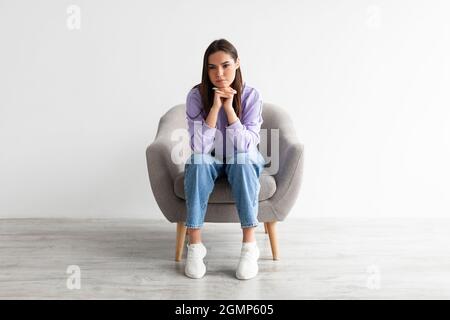 Die ganze Länge der jungen Frau, die in einem Sessel mit dumpfem oder nachdenklichem Gesichtsausdruck sitzt, Probleme oder Depressionen hat Stockfoto
