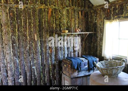 Holzstämme in einer Ecke, in einer Blockhütte. Ein Tisch mit einem Strohkorb. Sonnenlicht durch das Fenster. Stockfoto