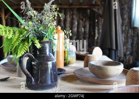 Nahaufnahme eines Keramikkännchens mit wilden Blumen, auf einem Tisch mit altem Geschirr aus Holz, in einer Blockhütte. Stockfoto