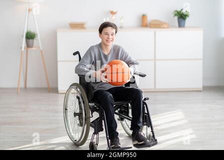 Fröhlicher behinderter Teenager, der Basketball hält, im Rollstuhl sitzt und drinnen die Kamera anlächelt Stockfoto