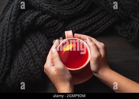 Hände halten Teetasse mit Zitrone auf dunklem Hintergrund. Schwarzer Strickschal im Hintergrund Stockfoto