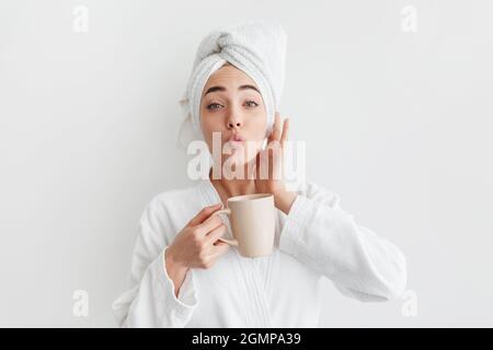 Lächelnd süße tausendjährige kaukasische blonde Dame im Bademantel und Handtuch auf dem Kopf, hält Tasse, trinkt Kaffee Stockfoto