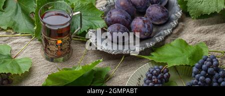 Trauben aus blauen, unvollkommenen Gartentrauben und Pflaumen mit Blättern in Vintage-Silberschale und schwarzem Tee aus antikem Glas Stockfoto