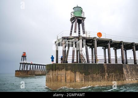 Whitby Hafen mit anonymen Männern, die während rauer See und Flut am Rand der Pier-Wand fischen. Farbenfrohe rote und grüne Leuchtturmtürme. Stockfoto