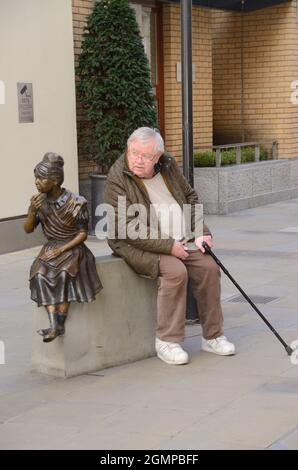 Der alte Mann saß neben der Statue des Mädchens auf einem Sockel. London, Großbritannien. Stockfoto