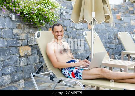 Mann mit Laptop, der sich auf der Sonnenliege am Pool entspannt Stockfoto