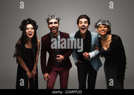 Aufgeregte multiethnische Freunde in halloween-Kostümen lachen vor der Kamera auf grau Stockfoto