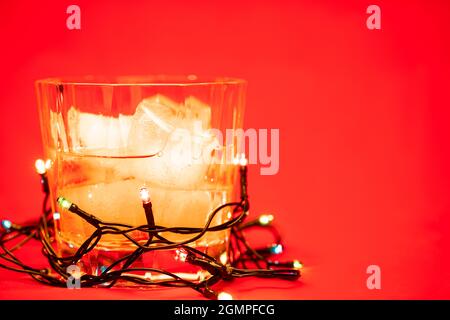 Nahaufnahme eines Glases Whiskey auf den Felsen mit weihnachtslichtern auf rotem Hintergrund - Konzept für weihnachtsgetränke Stockfoto