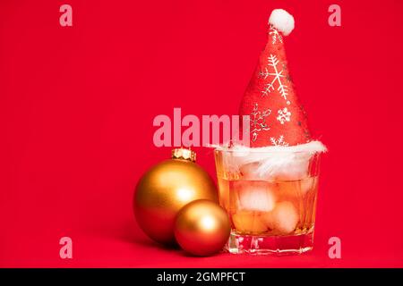 Bleiglas Whiskey auf den Felsen mit rotem weihnachtsmütze und weihnachtskugeln auf rotem Hintergrund - Weihnachtsgetränk-Konzept Stockfoto