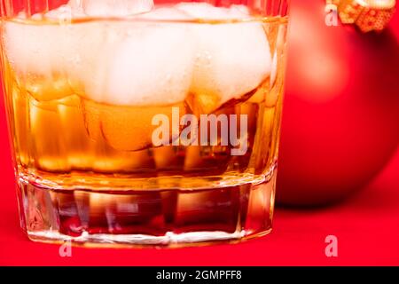 Nahaufnahme eines Whiskeyglases mit Eiswürfeln und Weihnachtskugel auf rotem Hintergrund - Weihnachtsgetränk-Konzept Stockfoto