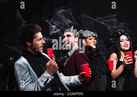 Fröhliche multiethnische Freunde mit Plastikbechern tanzen auf halloween-Party auf Schwarz Stockfoto