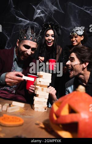Fröhlicher Mann in Vampir König Krone spielen Holzklötze Spiel während halloween-Party mit multiethnischen Freunden auf schwarz Stockfoto