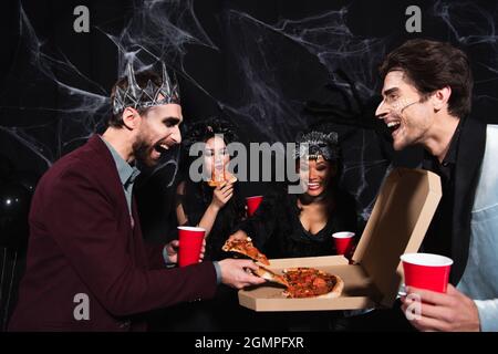 Glücklicher Mann in Vampirkönigskrone, der Pizza in der Nähe multiethnischer Freunde auf Schwarz nimmt Stockfoto