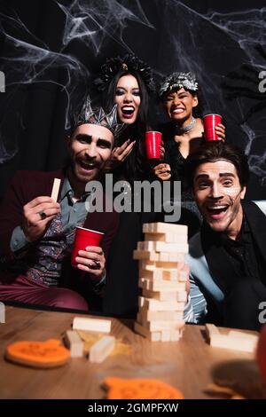 Aufgeregt multiethnischen Freunde in halloween-Kostüme Blick auf Kamera in der Nähe von Holzblöcken Spiel auf schwarz Stockfoto