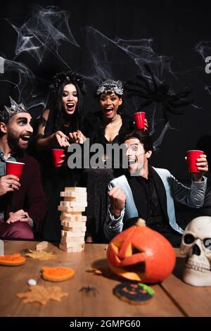 Aufgeregt Mann in halloween Make-up zeigt Win-Geste in der Nähe von Holzblöcken Spiel und multiethnischen Freunden auf schwarz Stockfoto
