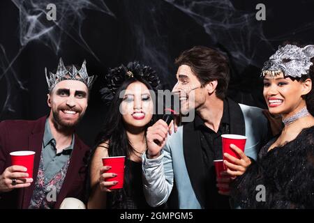 Mann in halloween Make-up singt Karaoke in der Nähe von multiethnischen Freunden mit Plastikbechern auf schwarz Stockfoto