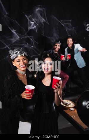 Glückliche interracial Frauen in halloween-Kostümen in der Nähe Mann winkende Hand auf verschwommenem und schwarzem Hintergrund Stockfoto