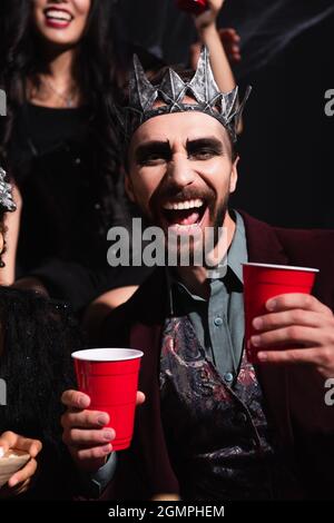 Mann in Vampir König Krone hält Kunststoff-Tasse in der Nähe verschwommen Frau auf schwarzem Hintergrund Stockfoto