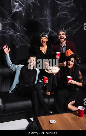 Mann winkt mit der Hand neben aufgeregten multiethnischen Freunden in halloween-Kostümen, die Popcorn essen und Bier auf Schwarz trinken Stockfoto