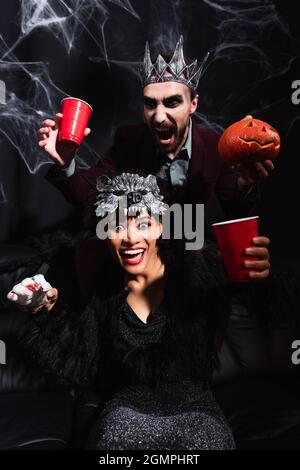 Gruselige afroamerikanische Frau in Werwolf-Maske hält Spielzeug Hand neben gruseligen Mann mit geschnitzten Kürbis auf schwarz Stockfoto