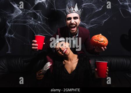 Lachender Mann im Vampir halloween Kostüm mit Plastikbecher und geschnitztem Kürbis bei afroamerikanischer Frau in Wolfmaske auf schwarz Stockfoto