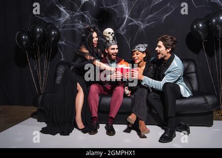 Multiethnische Freunde in halloween-Kostümen toasten mit Plastikbechern, während sie auf dem Sofa in der Nähe von Luftballons auf Schwarz sitzen Stockfoto