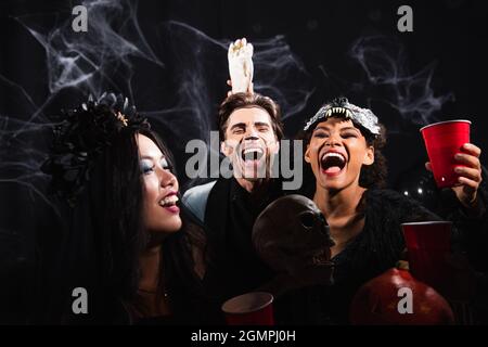 Lachende afroamerikanische Frau hält Spielzeughand in der Nähe aufgeregt Mann während halloween-Party auf schwarz Stockfoto
