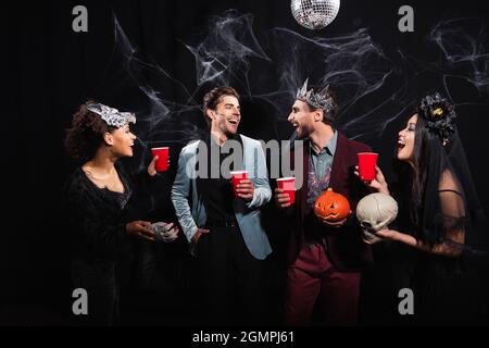 Glückliche multiethnische Freunde mit Plastikbechern reden während der halloween-Party auf Schwarz Stockfoto