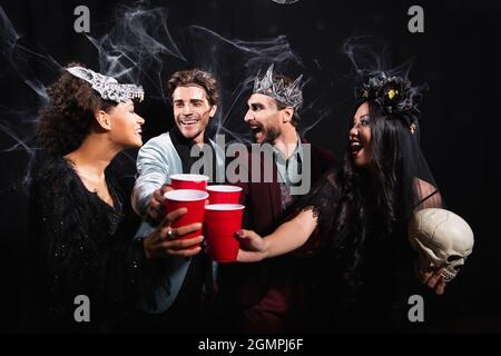 Aufgeregte multiethnische Freunde in halloween Kostümen toasten mit Plastikbechern auf Schwarz Stockfoto