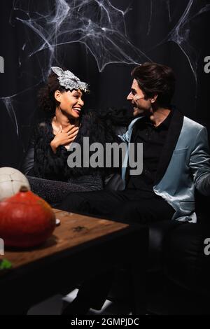 Lächelnde afroamerikanische Frau in Wolf halloween-Maske berühren Brust, während im Gespräch mit Mann auf schwarz Stockfoto