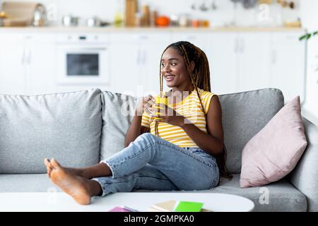 Entspannte afroamerikanische junge Frau, die auf der Couch mit Kaffeebecher chillt Stockfoto
