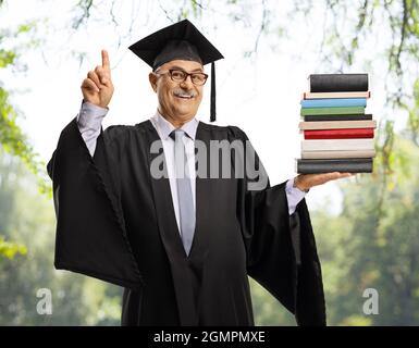 Glücklicher reifer Mann in einem Abschlusskleid, der einen Stapel Bücher hält und nach oben zeigt, mit Bäumen im Hintergrund Stockfoto