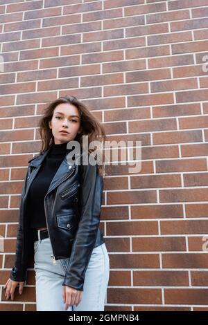 Hübsche junge Frau in schwarzer Lederjacke und Jeans, die in der Nähe der Ziegelmauer steht Stockfoto