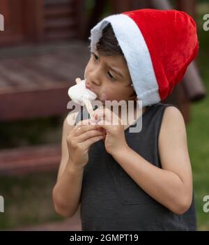 Lateinischer kleiner Junge im Profil, der zur Weihnachtszeit ein Popsicle isst, trägt einen roten Weihnachtsmann-Hut und ein ärmelloses T-Shirt. Vertikal. Argentinien Stockfoto