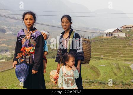 Sapa, Vietnam - 15. April 2016: Schwarze Hmong-Minderheitsfrau trägt Kind auf dem Rücken, während sie in den Bergen arbeitet und geht. Traditionelle Schlinge Stockfoto