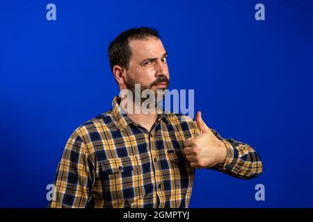 Unglücklicher, skeptischer, zweifelhafter Mann, der mit Daumen nach oben zeigt. Hipster-Männchen mit Bart in blau kariertem Hemd isoliert auf blauem Studio Stockfoto