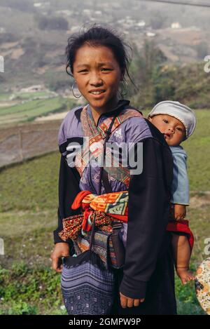 Sapa, Vietnam - 15. April 2016: Schwarze Hmong-Minderheitsfrau trägt Kind auf dem Rücken, während sie in den Bergen arbeitet und geht. Traditionelle Schlinge Stockfoto