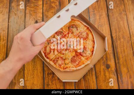 Die Hand des Typen öffnet eine Pappschachtel Pizza vor dem Hintergrund eines Holztisches. Köstliches Fast Food. Stockfoto