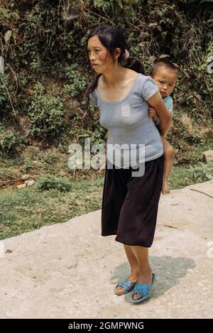 Sapa, Vietnam - 15. April 2016: Schwarze Hmong-Minderheitsfrau trägt Kind auf dem Rücken, während sie in den Bergen arbeitet und geht. Traditionelle Methode Stockfoto