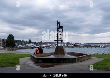 Kirkland, WA USA - ca. August 2021: Blick auf Menschen, die an einem Springbrunnen am Yachthafen sitzen, während Familien im Hintergrund am Strand spielen. Stockfoto