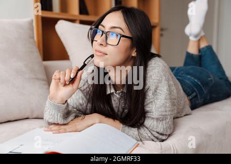 Zufrieden asiatische Student Mädchen tun Hausaufgaben während liegen auf Couch zu Hause Stockfoto